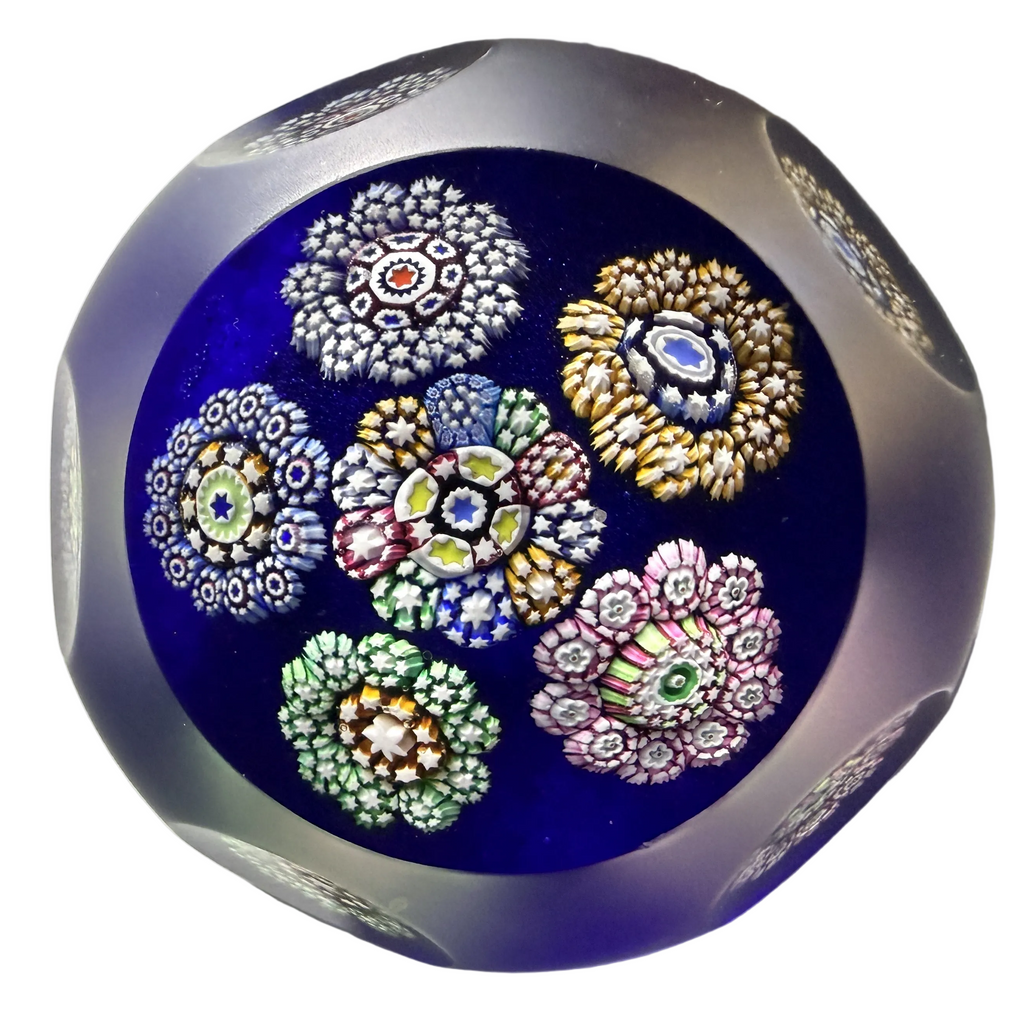 A round glass paperweight featuring a top facet and 5 side facets with 6 complex cane millefiori rondelles on top of translucent cobalt blue ground, sandblasted in between side facets.