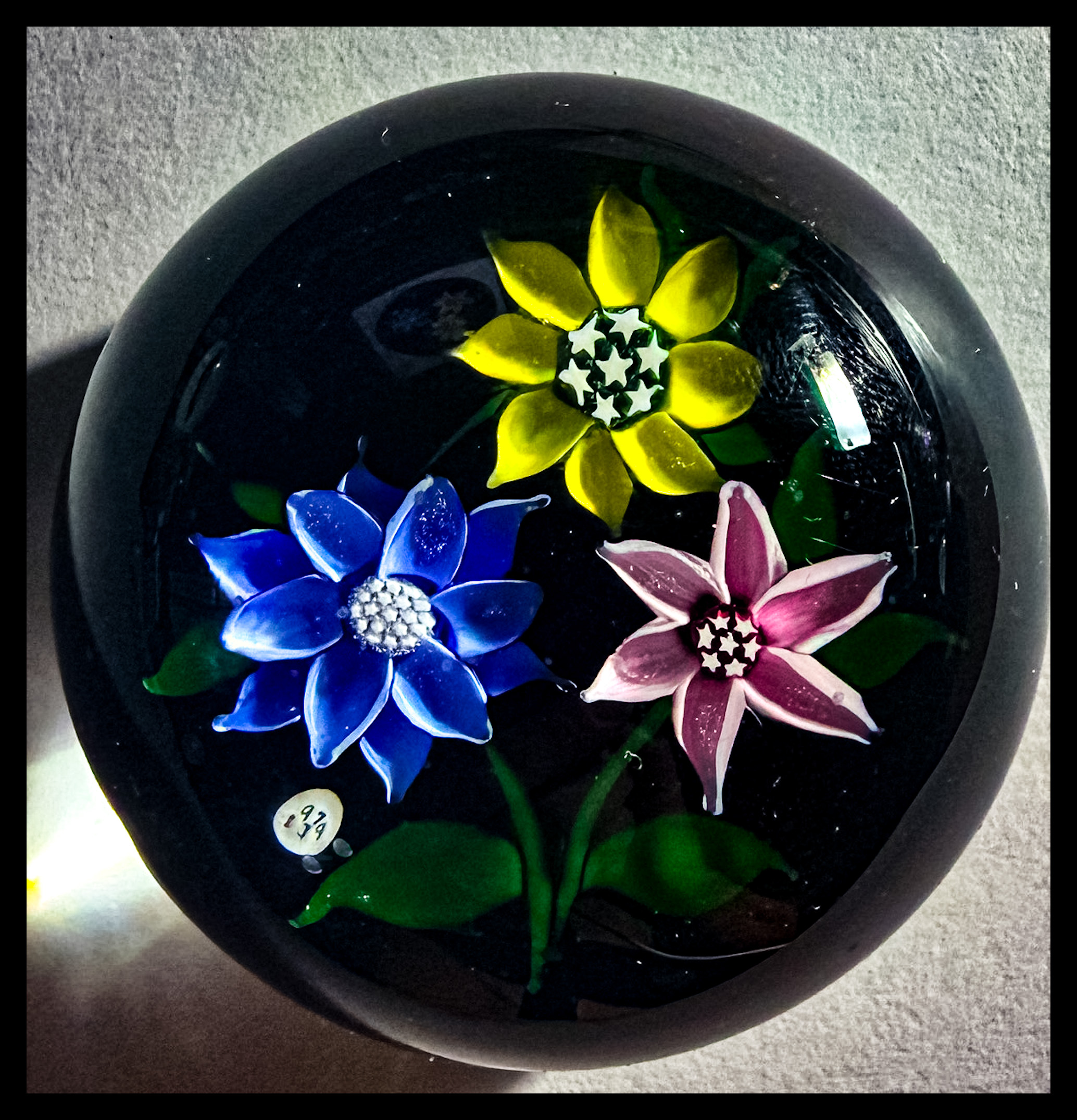 1979 J Glass Blue, yellow and pink flowerheads with stardust center canes atop an opaque black ground, J 1979 cane floats next to the blue flowerhead. Delicate lampwork at it;s finest with wonderfully cupped petals on each flower.&nbsp; A near perfect weight at a great price, easily one of the best among it's contemporaries in 1979.&nbsp; 2 1/4"