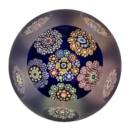 A round glass paperweight featuring a top facet and 5 side facets with 6 complex cane millefiori rondelles on top of translucent cobalt blue ground, sandblasted in between side facets.